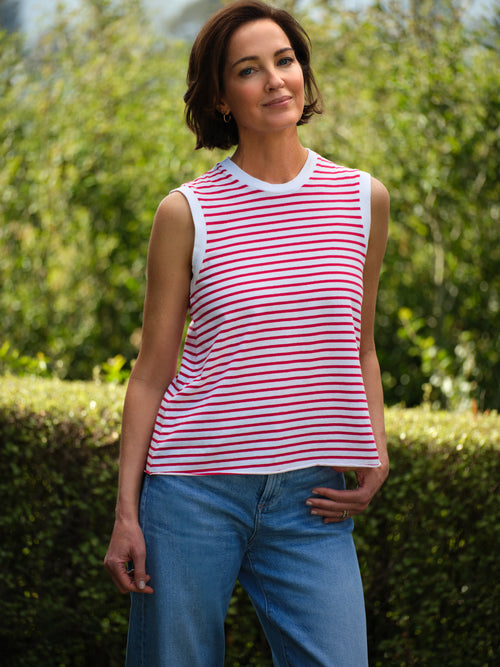 Woman wearing a red and white striped sleeveless top and blue jeans standing outdoors with greenery in the background., front view