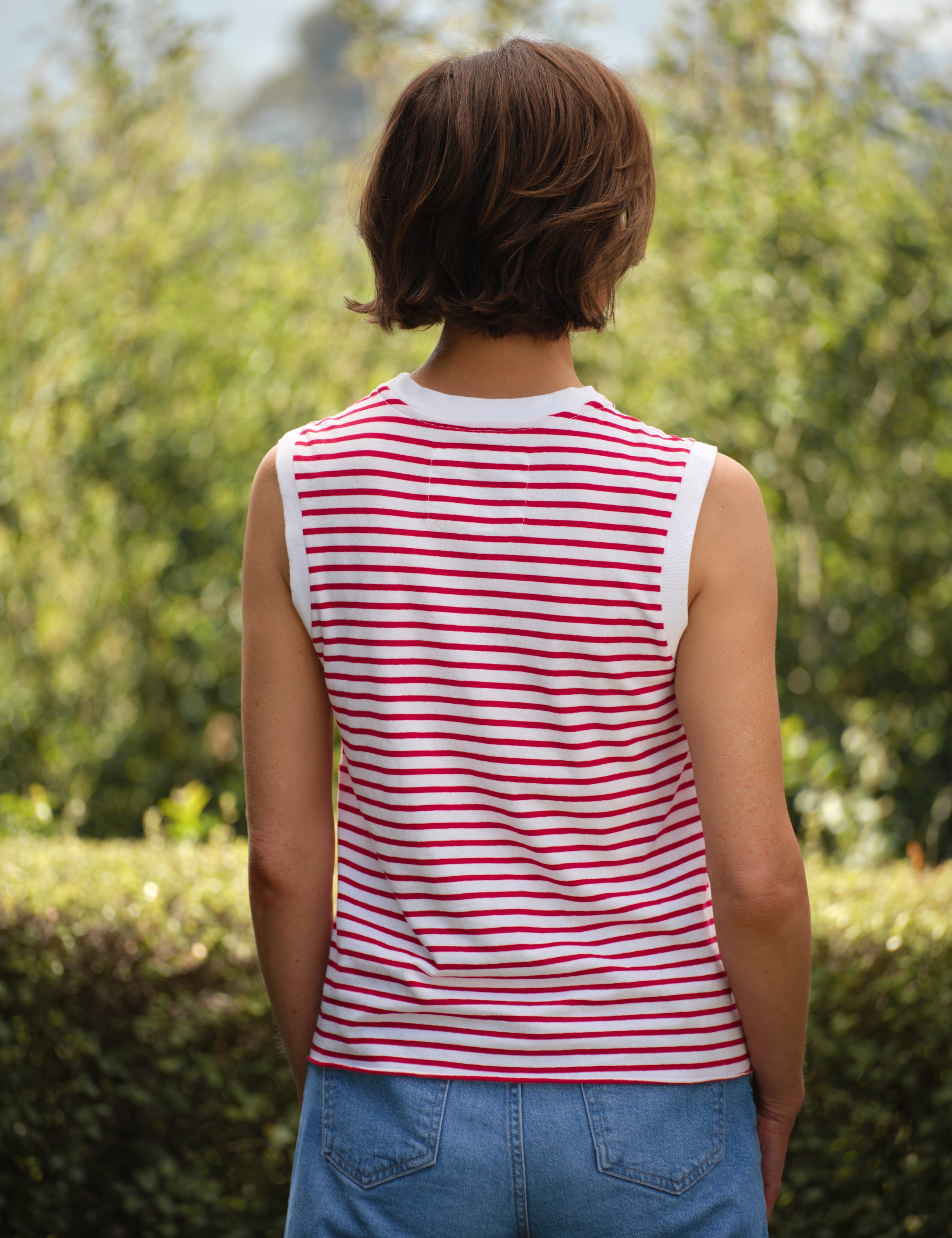 Person wearing a red and white striped sleeveless shirt with blue jeans, standing outdoors with greenery in the background., alternate view