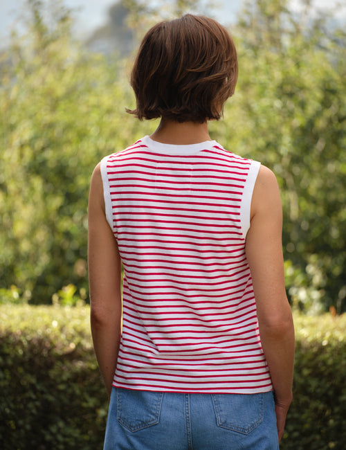 Person wearing a red and white striped sleeveless shirt with blue jeans, standing outdoors with greenery in the background., alternate view