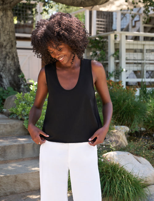 Woman wearing a black tank top and white pants standing outdoors with greenery and a building in the background., front view