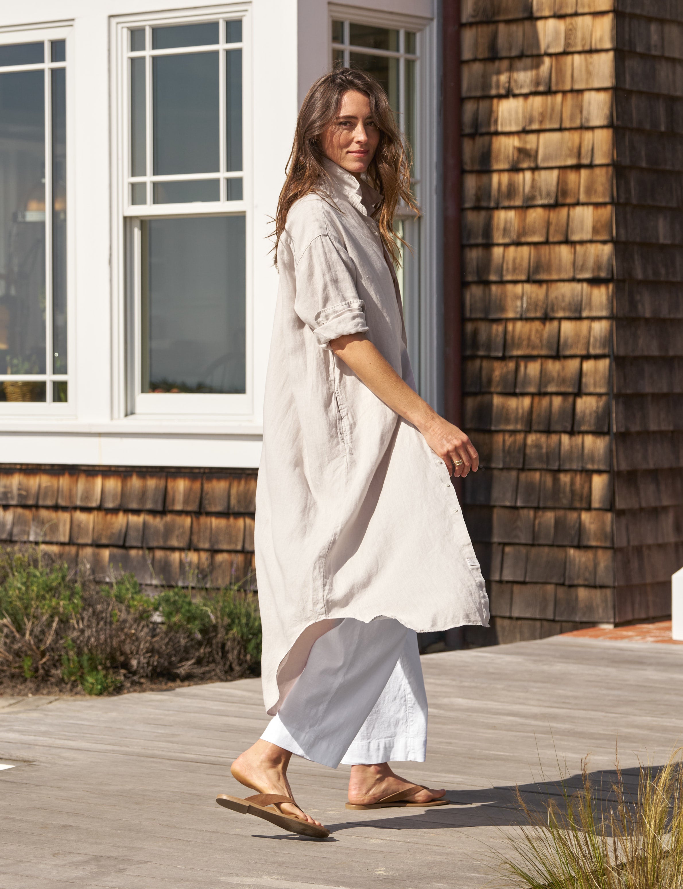 Woman in a long, light-colored dress standing on a wooden deck next to a wooden building., alternate view