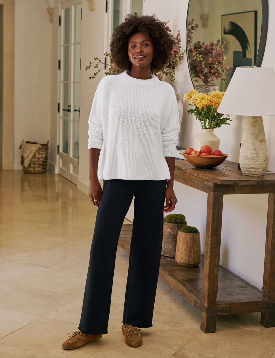 Woman in a white sweater and navy pants standing in a room with wooden flooring and a wooden console table.