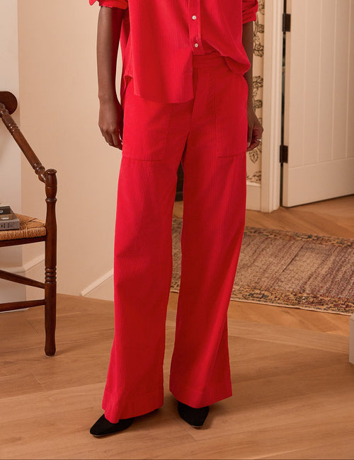 A woman stands in a warmly lit indoor room wearing a bright red matching two-piece outfit consisting of a button-front collared shirt and wide-leg pants., view 4