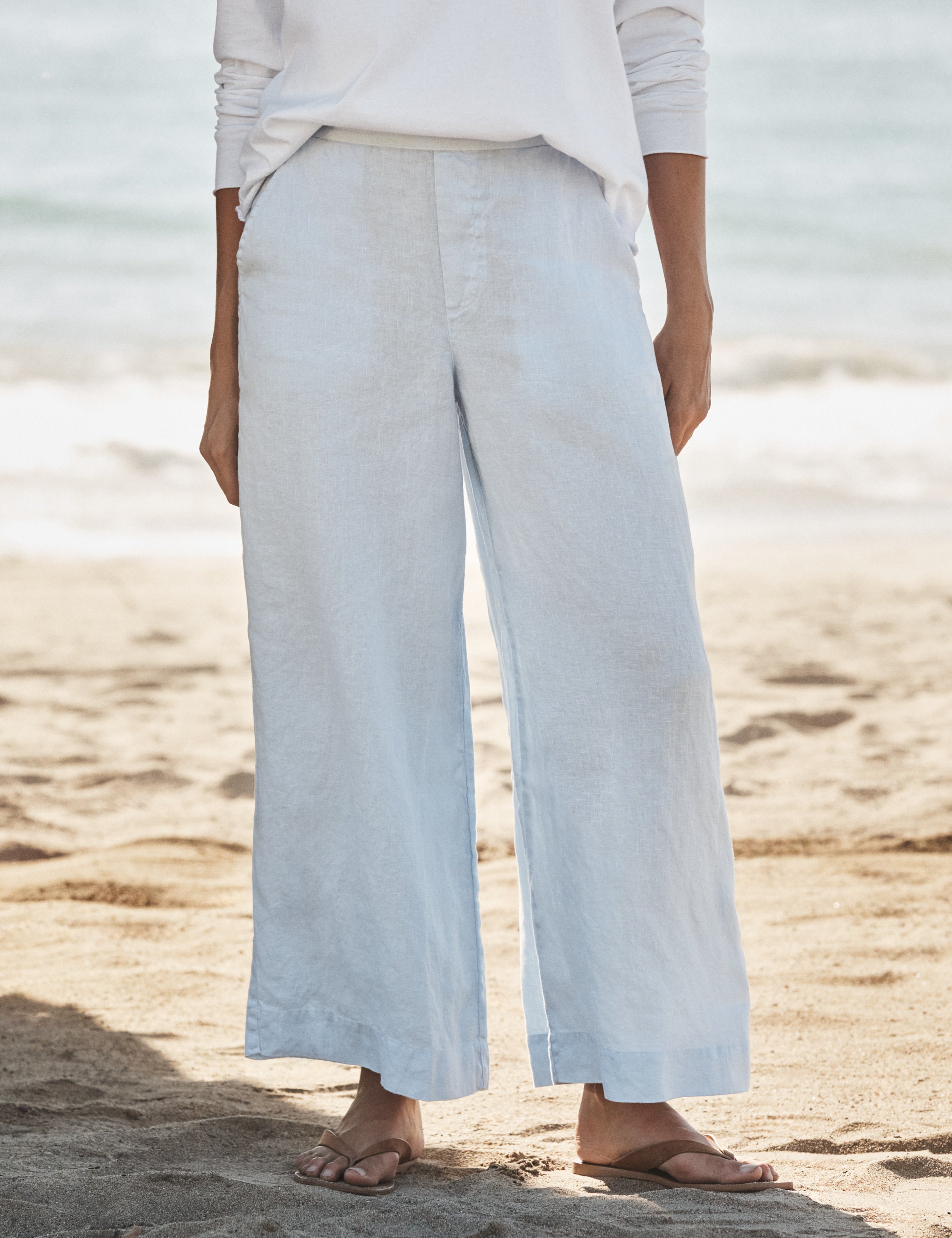 Person wearing light blue wide-leg pants and a white top on a sandy beach.