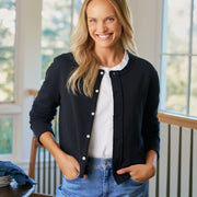 Woman wearing a black cardigan, white shirt, and blue jeans indoors.