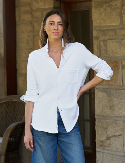 A woman wearing a white long-sleeve button-up shirt with rolled cuffs and blue jeans, standing in front of a stone wall., front view