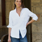 A woman wearing a white long-sleeve button-up shirt with rolled cuffs and blue jeans, standing in front of a stone wall.