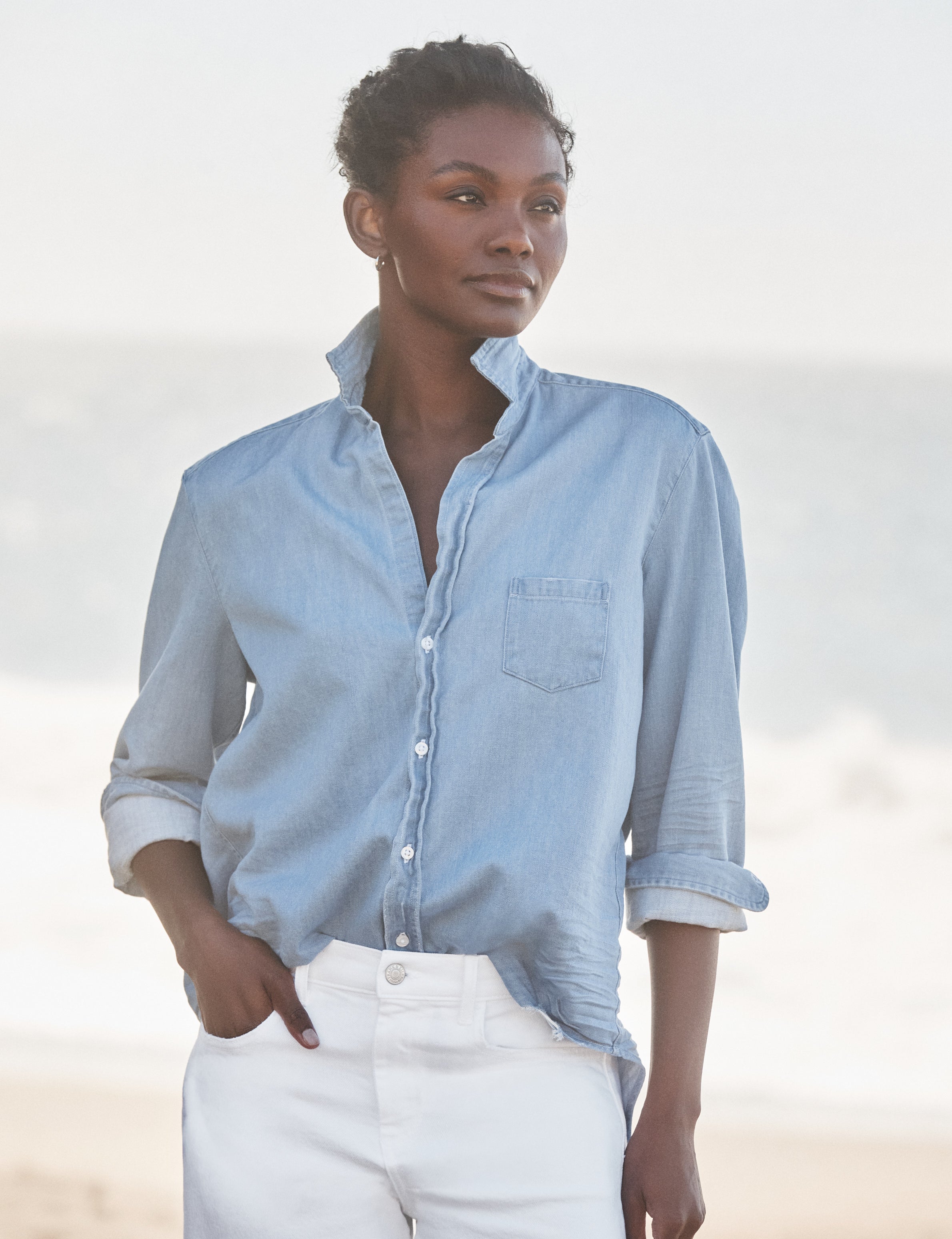 Woman wearing a light blue button-up shirt and white pants against a blurred natural background, view 4