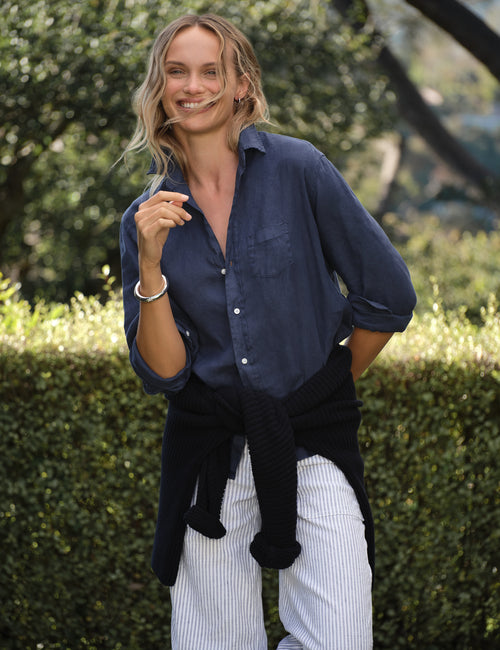 A woman standing outdoors in front of a green hedge, wearing a dark blue button-up shirt, white and blue vertically striped pants, and a navy knit sweater tied around her waist., view 4