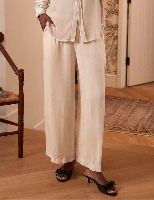 A woman modeling a matching cream-colored two-piece lounge set consisting of a long-sleeve button-up shirt and wide-leg pants, standing in a warmly lit room with wood floors and a wooden chair., front view