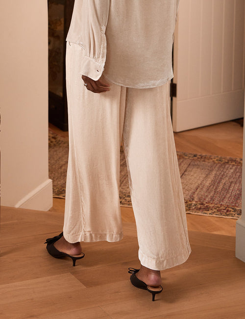 A woman viewed from behind wearing a cream-colored, long-sleeve button-up shirt and matching wide-leg trousers, standing in a home interior with wood floors and a doorway., alternate view