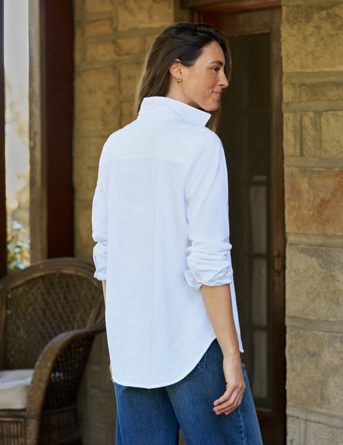 A woman wearing a white long-sleeve button-up shirt and blue jeans, standing on a stone porch next to a wicker chair., alternate view
