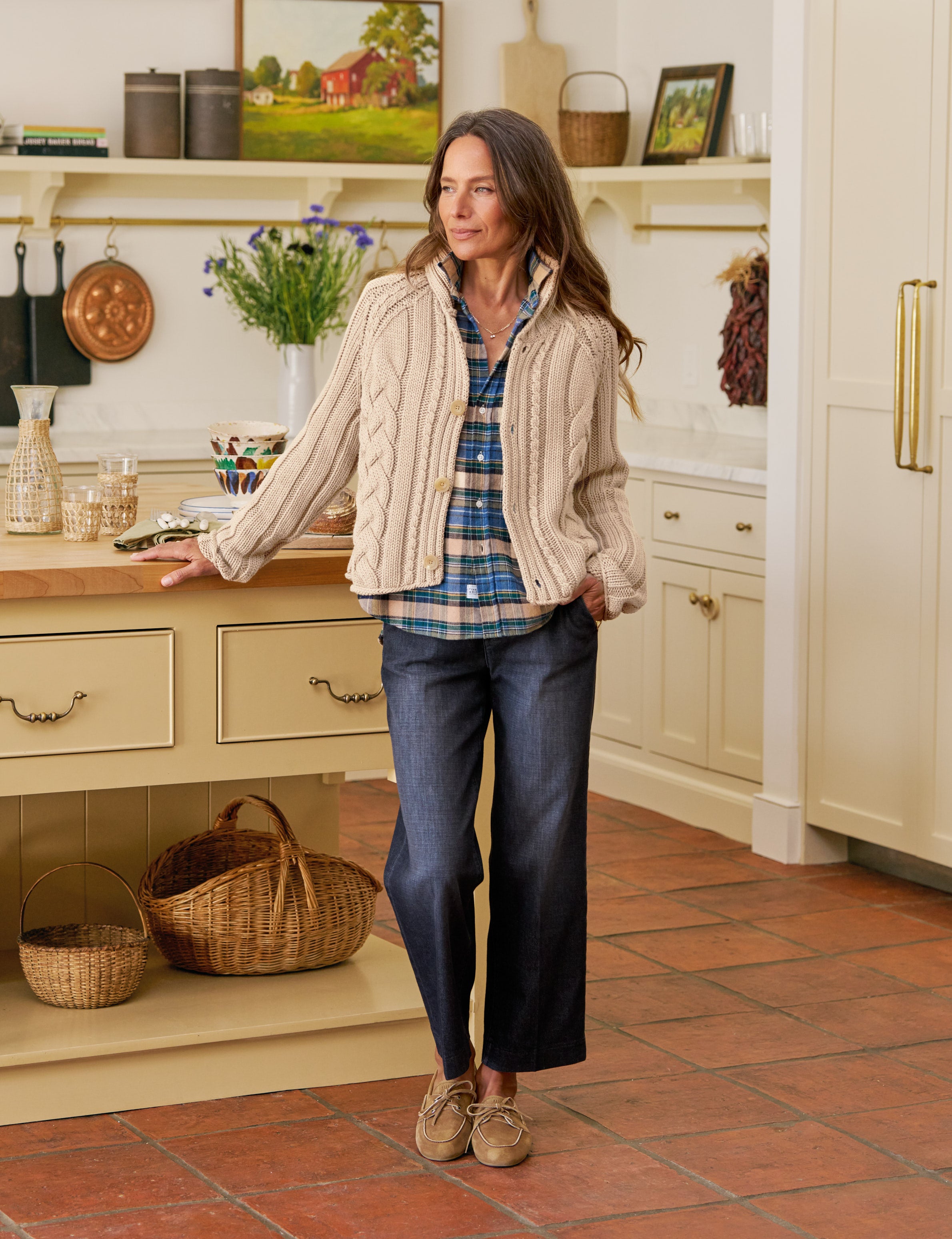 Woman standing in a kitchen wearing a plaid shirt, beige cardigan, and blue jeans.
