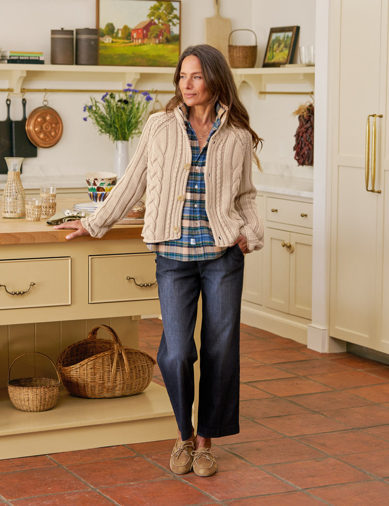 Woman standing in a kitchen wearing a plaid shirt, beige cardigan, and blue jeans.
