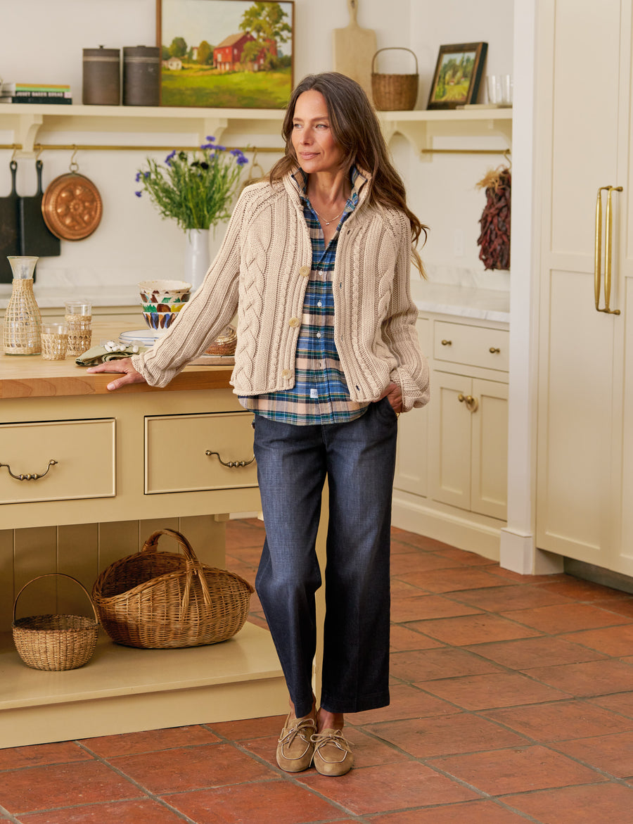 Woman standing in a kitchen wearing a plaid shirt, beige cardigan, and blue jeans.