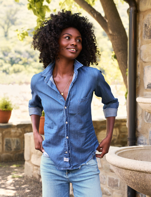 A woman with voluminous dark curly hair wearing a light blue chambray button-down shirt and light-wash jeans, standing outdoors in front of a stone wall and garden trees., front view