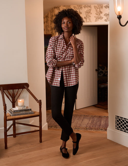 A woman stands in a warmly lit indoor room wearing a red and white checkered button-down shirt, black pants, and black flats. A wooden chair with a lit candle sits to the left, and an open doorway and patterned wallpaper are visible in the background., view 4