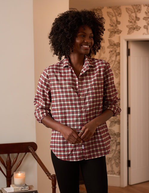 A woman with curly black hair wearing a red and white gingham button-down shirt and black pants, standing in a warmly lit room with patterned wallpaper., front view