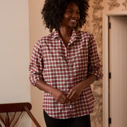 A woman with curly black hair wearing a red and white gingham button-down shirt and black pants, standing in a warmly lit room with patterned wallpaper.