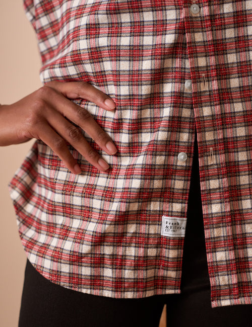 Close-up of a person wearing a red, white, and black plaid button-up shirt, with a hand resting on the fabric and a small sewn label near the hem., view 3