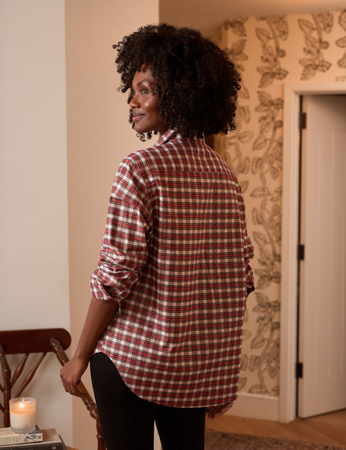 A woman with curly black hair is shown from the back wearing a red and white plaid button-down shirt and black pants in an indoor setting with patterned wallpaper., alternate view