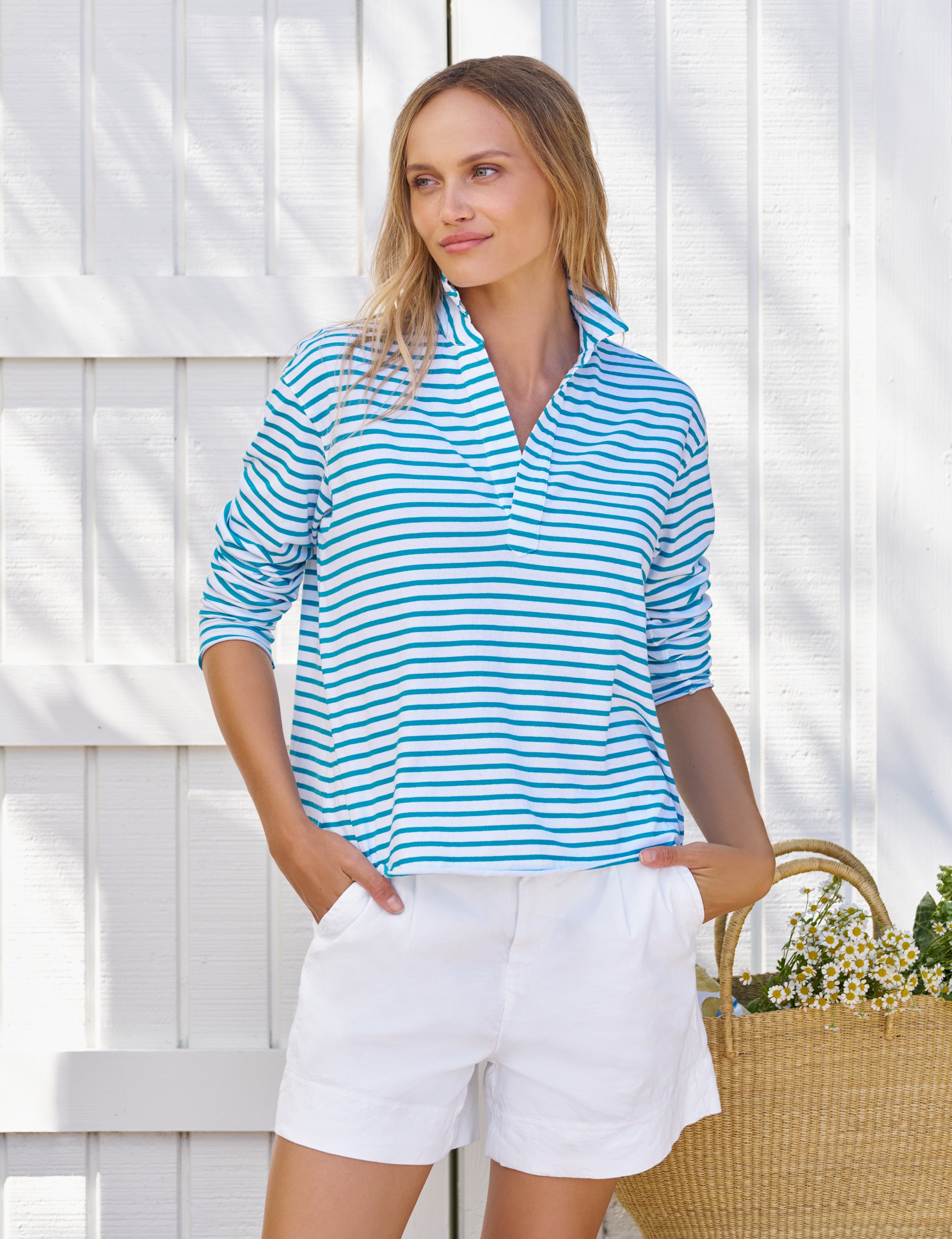 Woman wearing a blue and white striped shirt and white shorts standing against a white wooden fence., front view