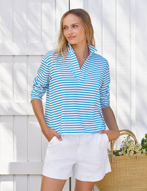 Woman wearing a blue and white striped shirt and white shorts standing against a white wooden fence., front view