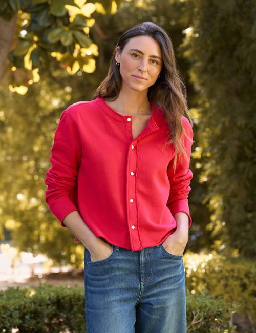 A woman wearing a bright red long-sleeve button-up shirt and blue jeans, standing outdoors in front of a blurred green garden background., front view