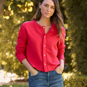 A woman wearing a bright red long-sleeve button-up shirt and blue jeans, standing outdoors in front of a blurred green garden background.