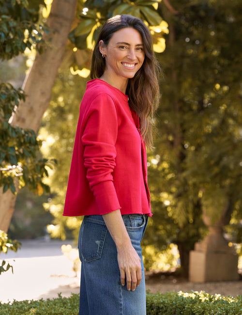 A woman wearing a bright red cropped long-sleeve top and blue jeans, standing outdoors in front of a softly blurred background of trees and foliage., alternate view