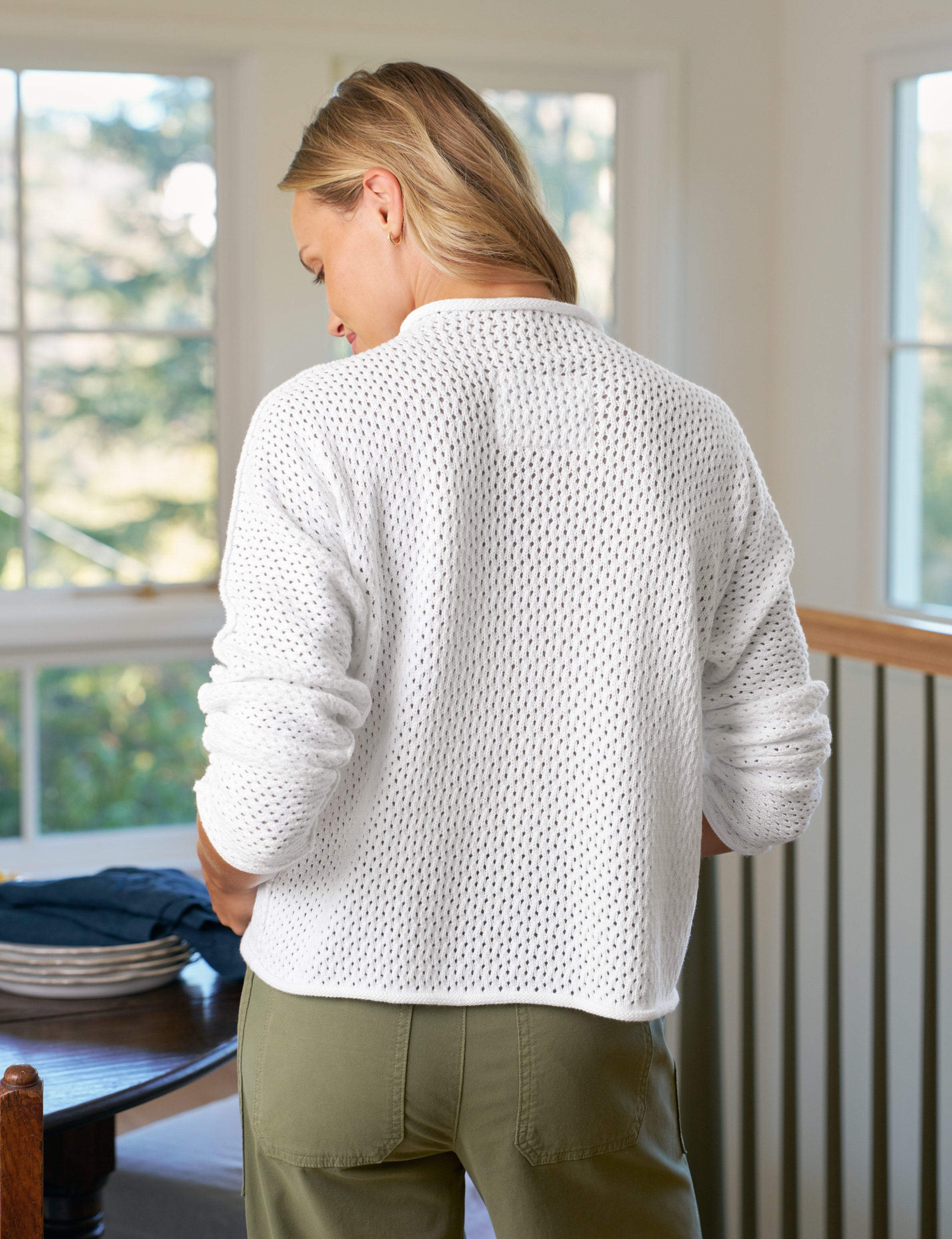 Person wearing a white textured sweater and green pants standing in a room with large windows.