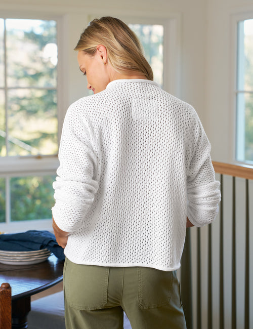 Person wearing a white textured sweater and green pants standing in a room with large windows., alternate view