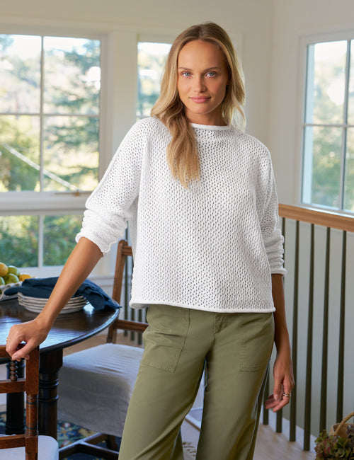 Woman wearing a white sweater and green pants standing in a room with large windows., front view