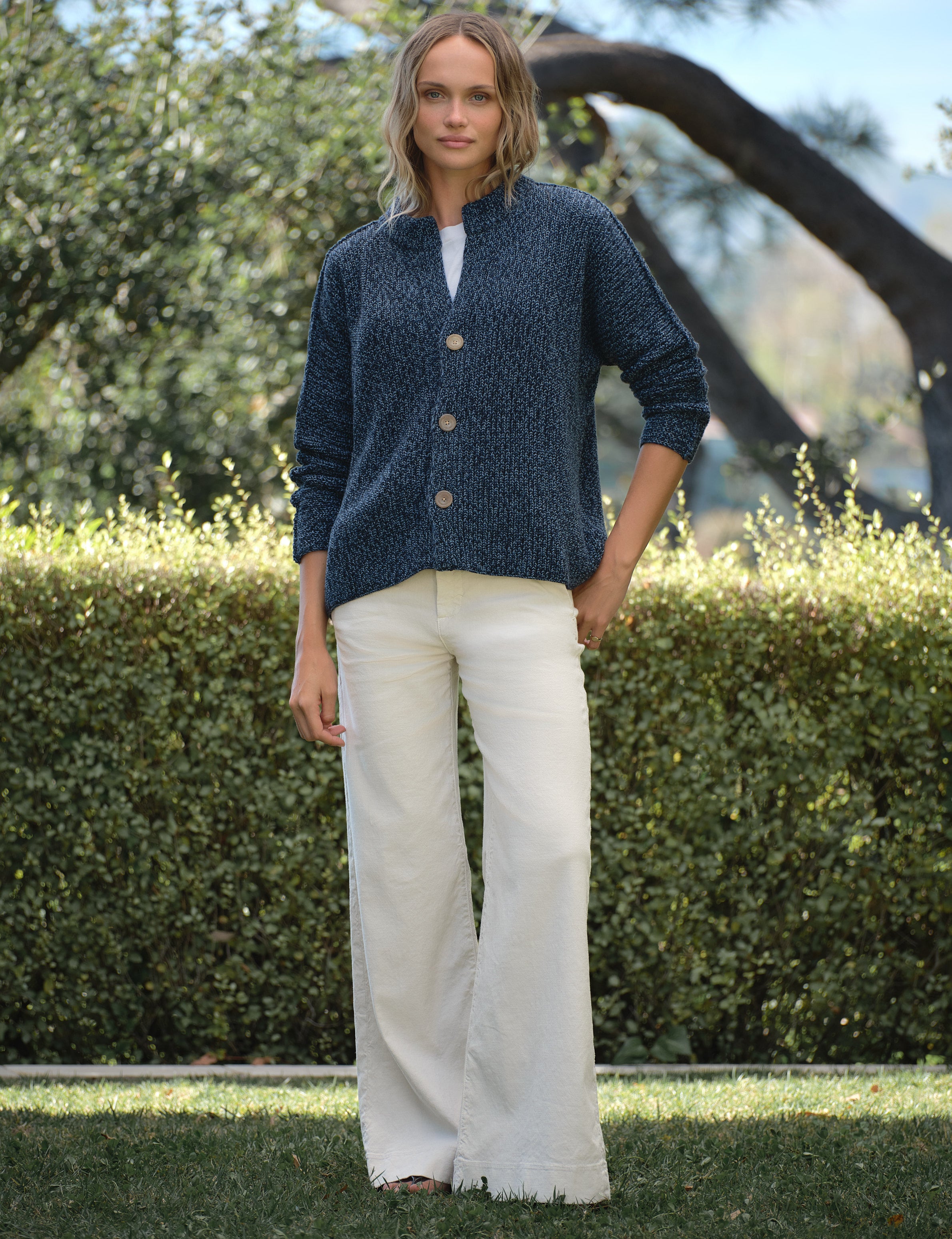 Woman wearing a blue cardigan and white pants standing outdoors with greenery in the background, view 3