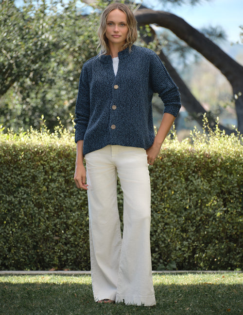 Woman wearing a blue cardigan and white pants standing outdoors with greenery in the background, view 3