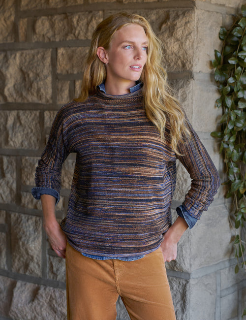 A woman wearing a textured, horizontally striped knit sweater in navy, blue, and brown tones, layered over a blue collared shirt and paired with tan pants, standing against a rustic stone wall with green ivy., front view
