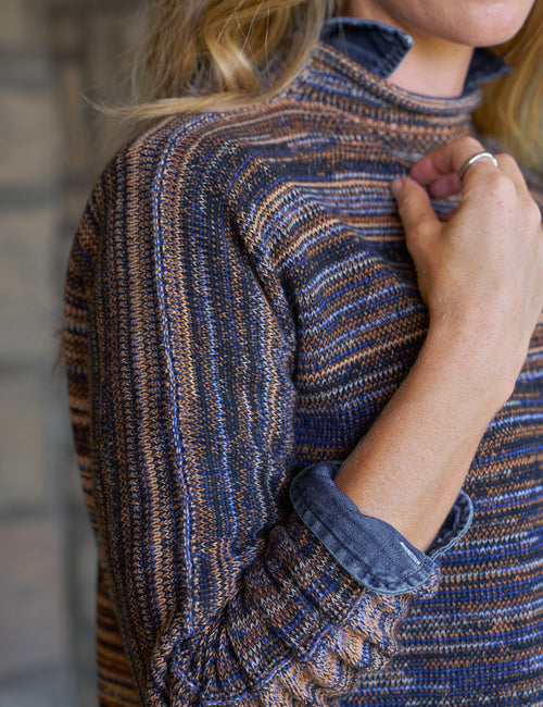 Close-up of a person wearing a textured knit mock-neck sweater with horizontal stripes in navy blue, brown, and tan, with a rolled-up sleeve showing a denim button cuff., view 3