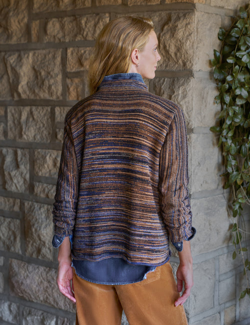A woman shown from the back wearing a textured, striped knit cardigan in brown, blue, and orange tones, layered over a blue shirt and paired with brown pants, standing against a stone wall., alternate view