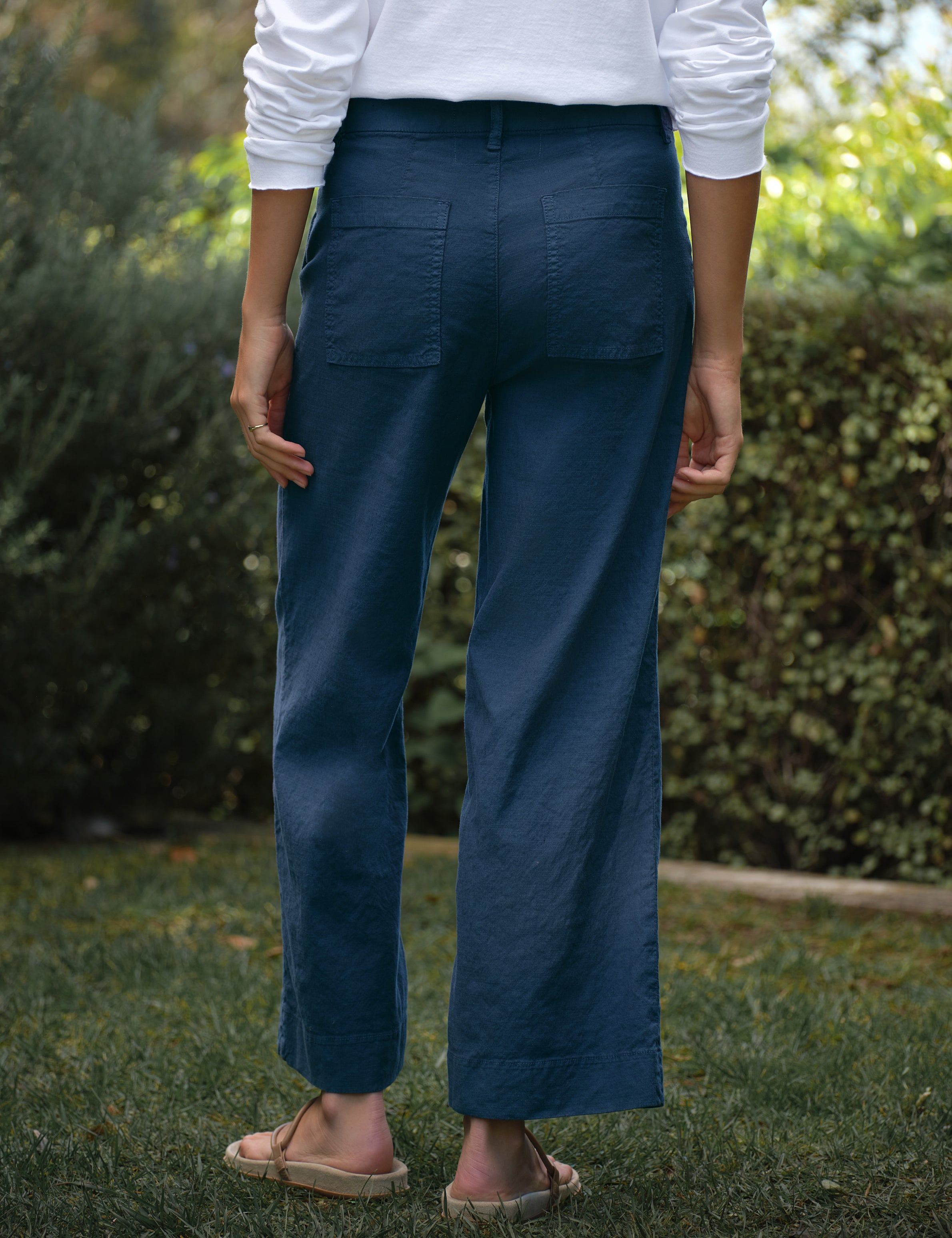 Person wearing blue pants and a white shirt standing outdoors with greenery in the background, alternate view
