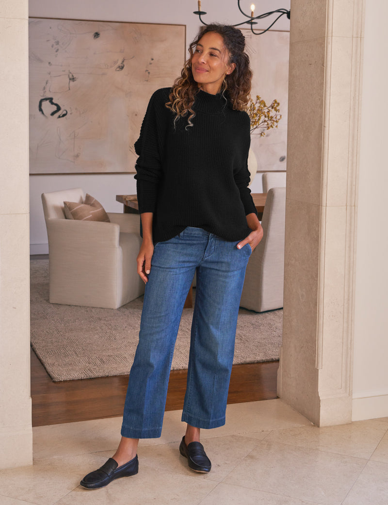 Woman wearing a black sweater and blue jeans standing in a room with a couch and artwork.