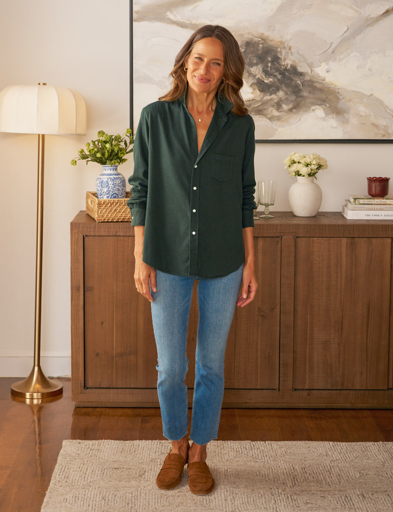 Woman wearing a green shirt and blue jeans standing in a room with wooden furniture and decorative items.