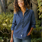 A woman wearing a blue denim button-up shirt and jeans, standing outdoors in front of green foliage and a stone wall.