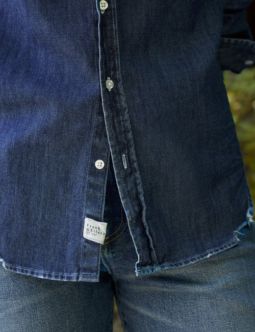 Close-up of a dark indigo denim button-up shirt worn with matching blue jeans, showing the fabric texture, silver buttons, and a small white brand label., view 3