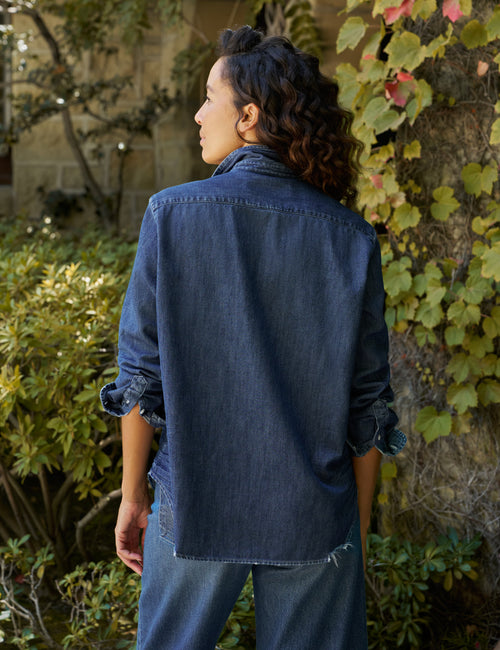 A woman wearing a dark blue denim shirt and jeans, photographed from behind in an outdoor garden setting with greenery and a tree trunk., alternate view