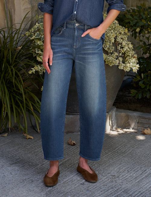 A person wearing medium-wash blue denim jeans with a barrel leg fit, paired with a blue denim shirt and brown suede flats, standing outdoors on a stone patio with potted plants in the background., front view