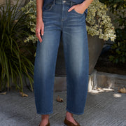 A person wearing medium-wash blue denim jeans with a barrel leg fit, paired with a blue denim shirt and brown suede flats, standing outdoors on a stone patio with potted plants in the background.