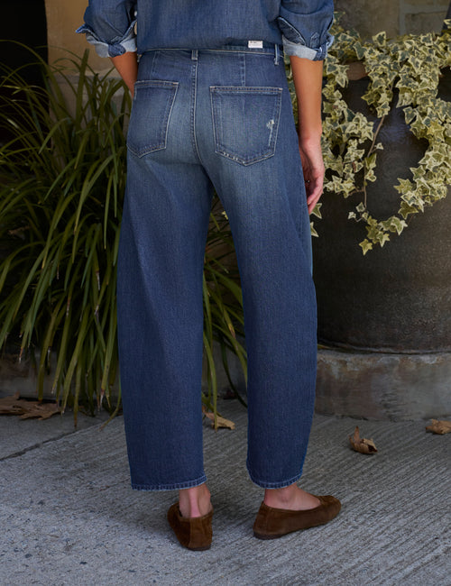 Rear view of a person wearing barrel-leg blue denim jeans with a button fly, paired with brown suede flats, standing on a concrete surface with potted plants in the background., alternate view