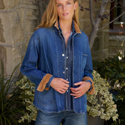 A woman wearing a blue denim button-up shirt with tan corduroy collar and cuffs, paired with blue jeans, standing outdoors in front of a stone wall and plants.