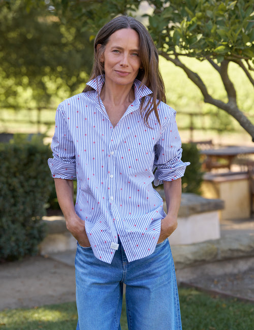 Woman wearing a striped shirt and jeans standing outdoors with greenery in the background, front view