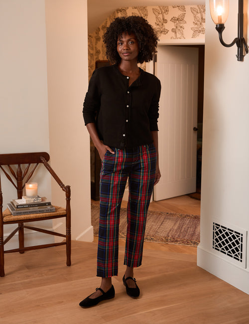 A woman stands in a home interior wearing a black long-sleeve button-up shirt and red, green, and navy plaid cropped pants with black flats., view 4